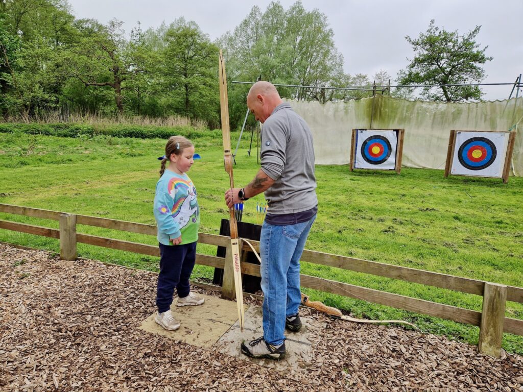 7 year old girl stood with a bald man, who is showing her how to hold an archery bow