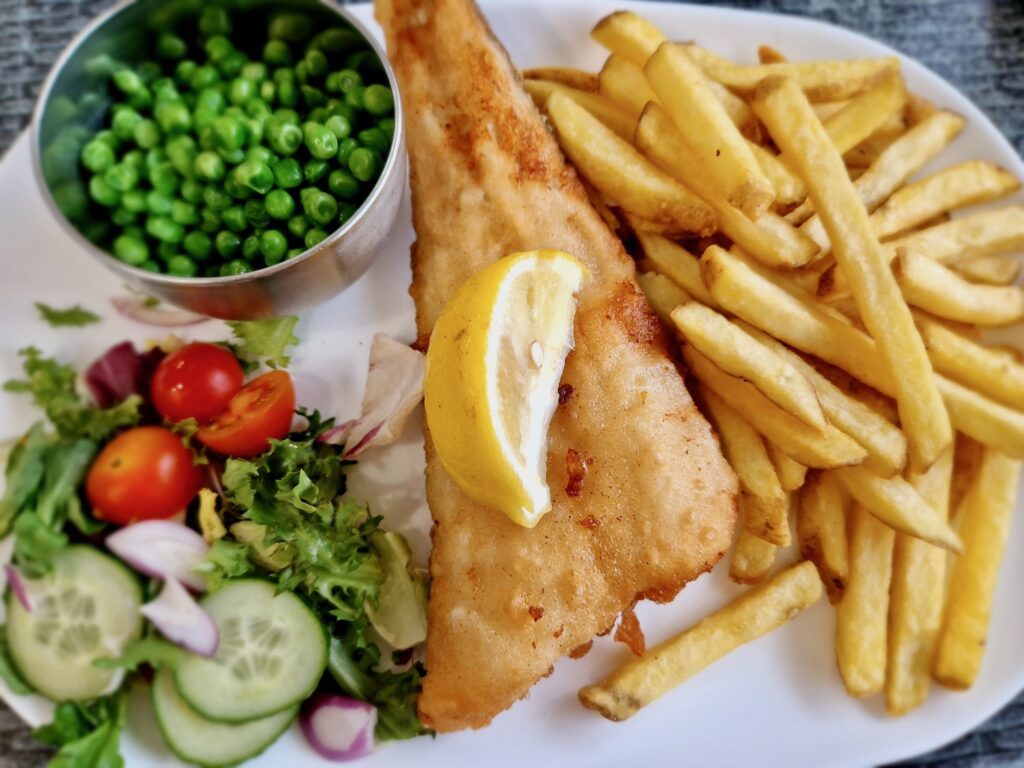 Fish and chips at Sara's Tearooms. A plate with chips, battered fish with a lemon on top, salad and a pot of peas