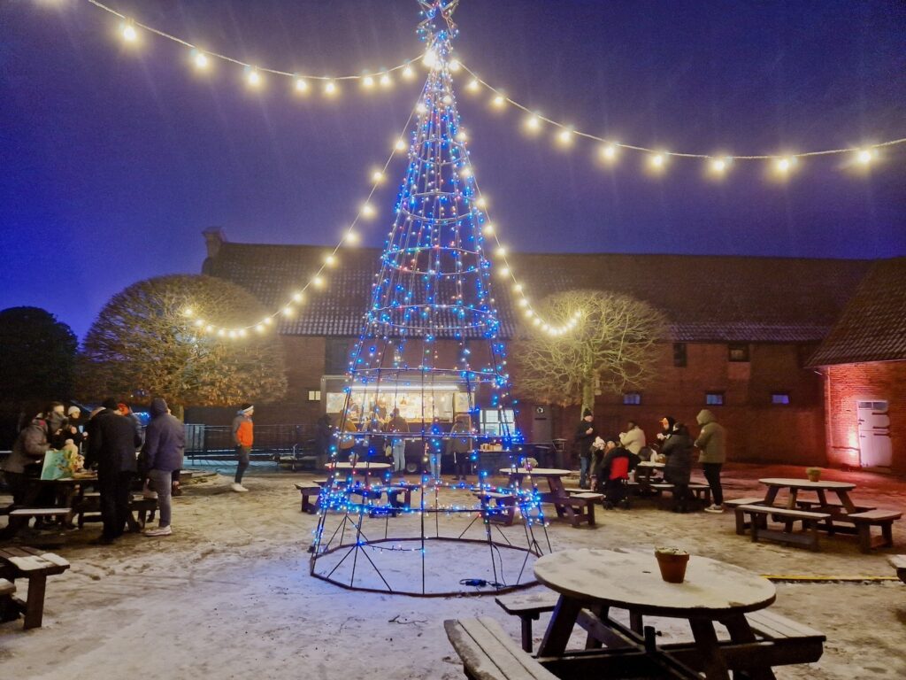 Blickling Wild Winter After Dark festive farmyard. A view of a lit up Christmas tree with a food stall and people in the background