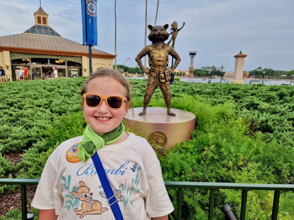 Erin, a 7 year old girl, wearing a cooling towel and sunglasses at Epcot. She is stood in front of a statue of Rocket and Groot from Guardians of the Galaxy