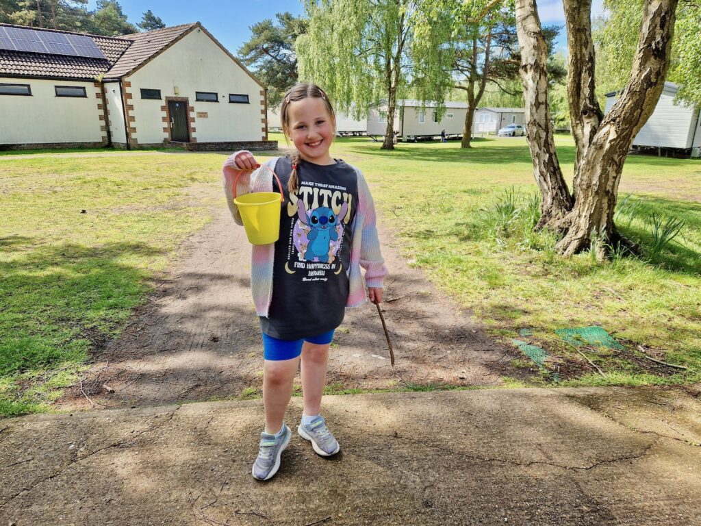 An 8 year old girl wearing a bright cardigan, blue shorts and a Lil and Stitch t-shirt, holding a yellow bucket up and a stick in the other hand