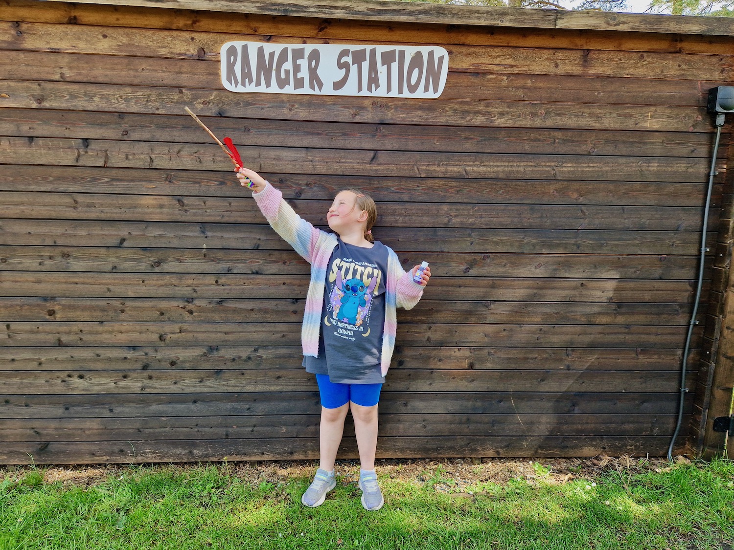 An 8 year old girl wearing a bright cardigan, blue shorts and a Lilo and Stitch t-shirt, holding up a magic wand made from a stick, with a potion bottle in the other hand