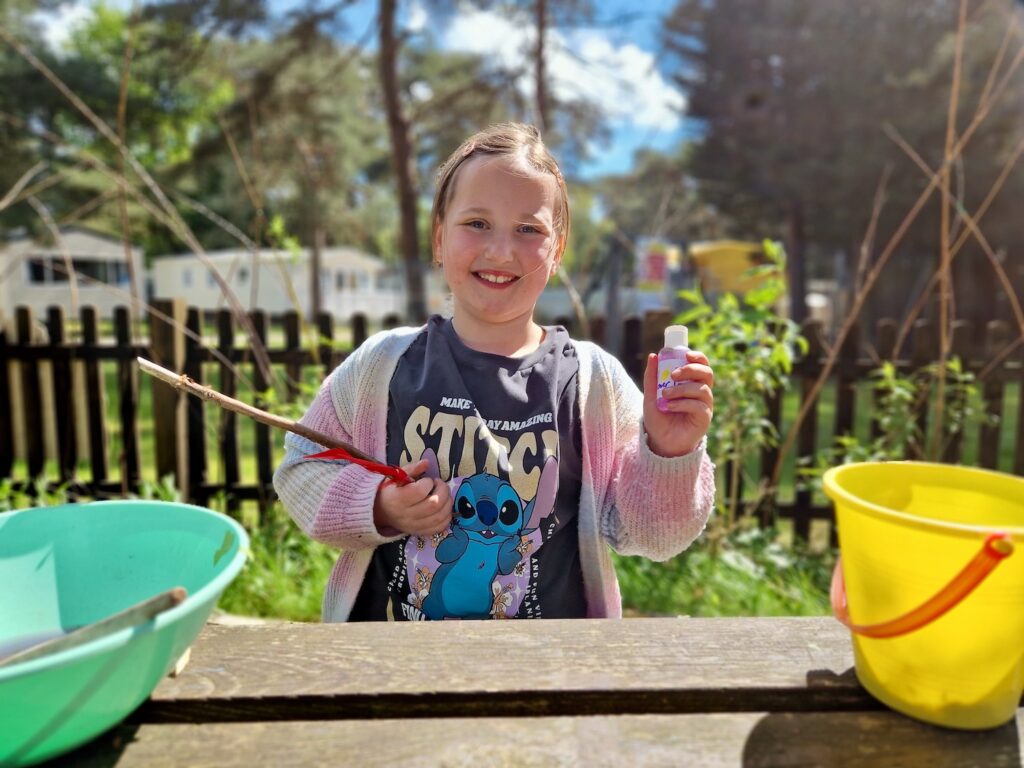 An 8 year old girl wearing a bright cardigan, blue shorts and a Lilo and Stitch t-shirt, holding a potion bottle and a wand made from a stick in the other hand