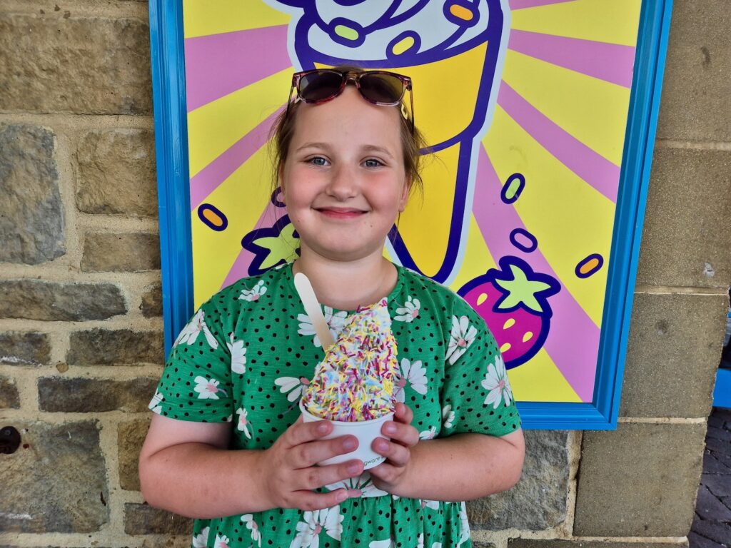 Erin, an 8 year old girl in a bright green floral dress and sunglasses on her head with a tub of ice cream with sprinkles from the Ice Cream Parlour at Legoland Windsor