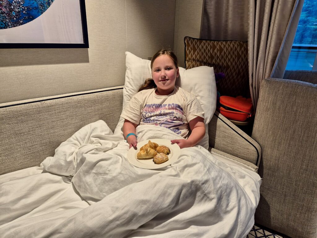 A young girl sits comfortably in bed in a P&O Iona balcony cabin, enjoying breakfast with croissants on a plate, with curtains partially open revealing the scenic fjords.
