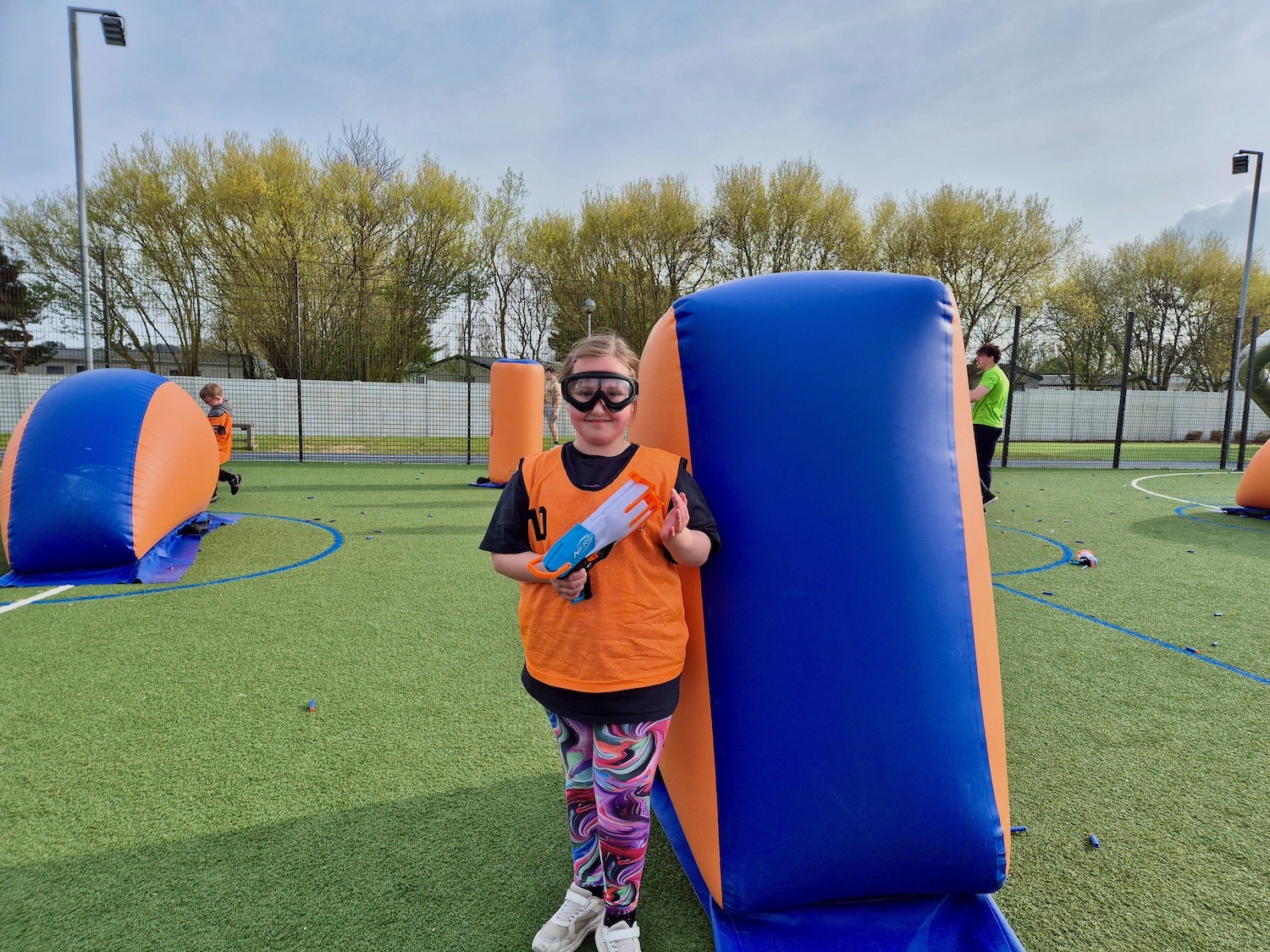 Child wearing goggles and orange vest with a Nerf blaster, standing on an inflatable obstacle course.