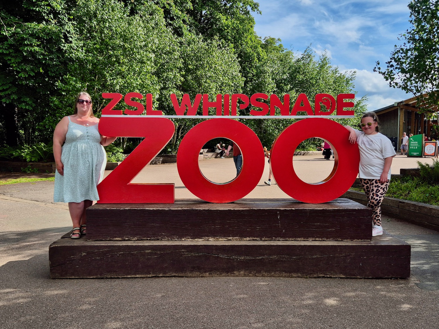Woman and girl standing on either side of the large red ZSL Whipsnade Zoo sign at the entrance to the zoo.