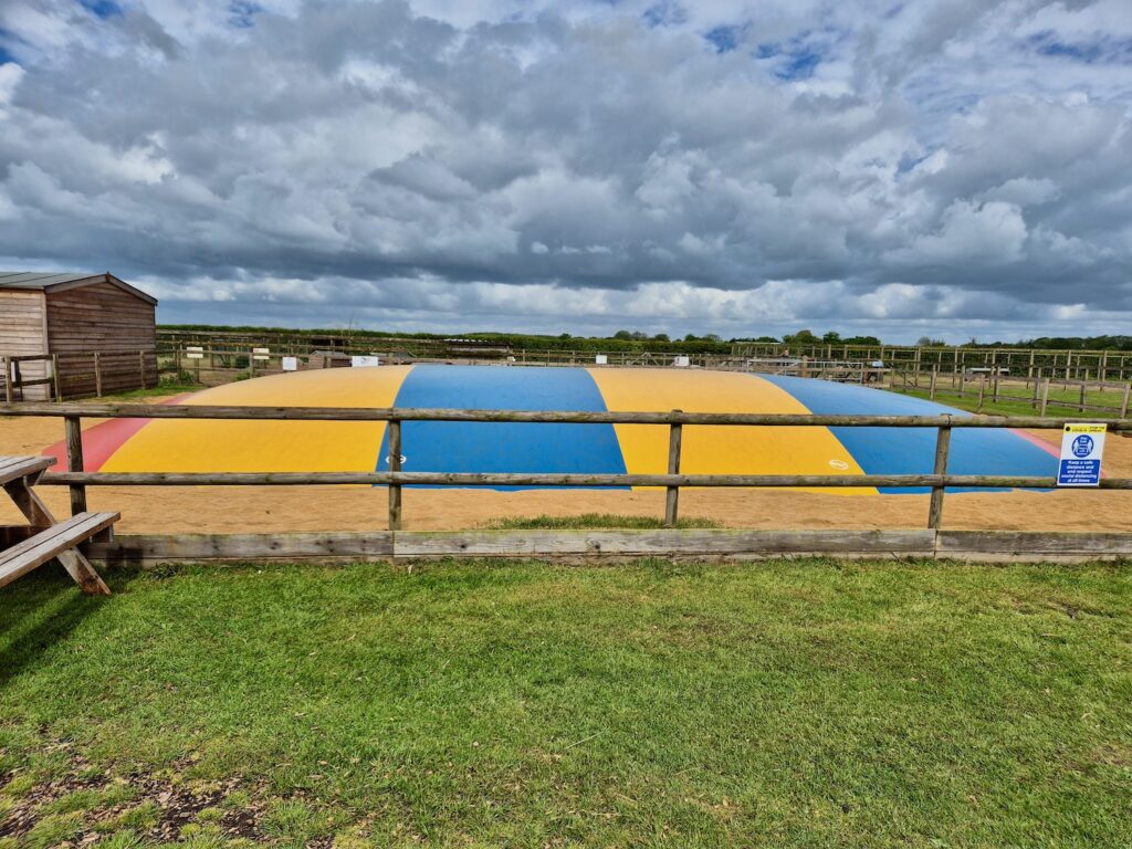 A large outdoor inflatable jumping pillow in red, yellow and blue at Camel Park Oasis, surrounded by grass and fencing.