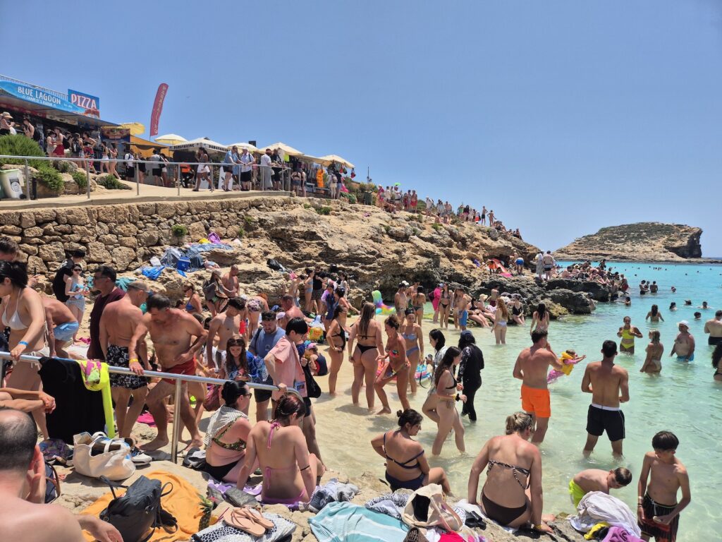 Crowded beach and rocky shoreline at the Blue Lagoon in Comino, Malta, with people sunbathing and swimming