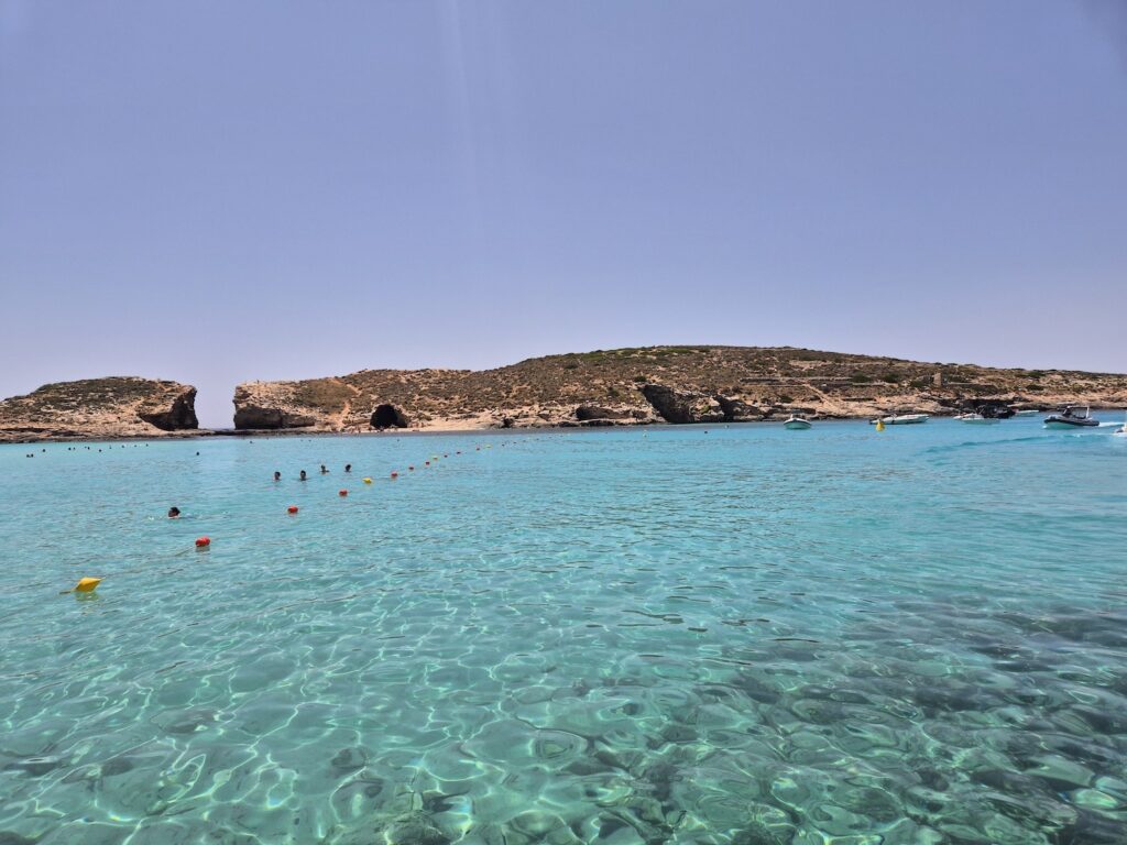 Clear turquoise waters and rocky coastline of the Blue Lagoon in Comino, Malta, with swimmers and boats in the distance