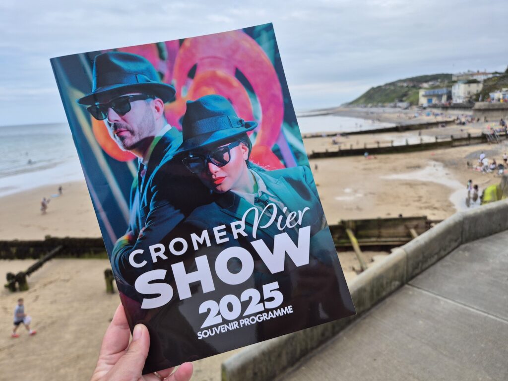 Hand holding the 2025 Cromer Pier Show souvenir programme with Cromer beach in the background