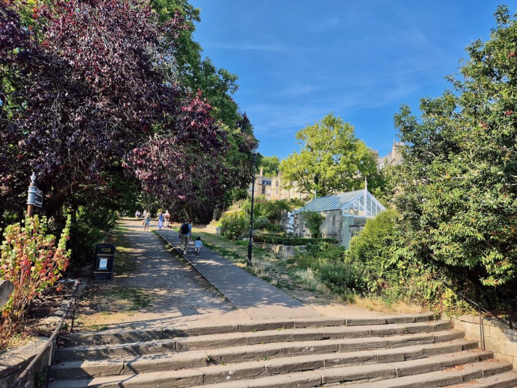A path winding up a hill in a park, surrounded by trees and a glasshouse with steps in the foreground.