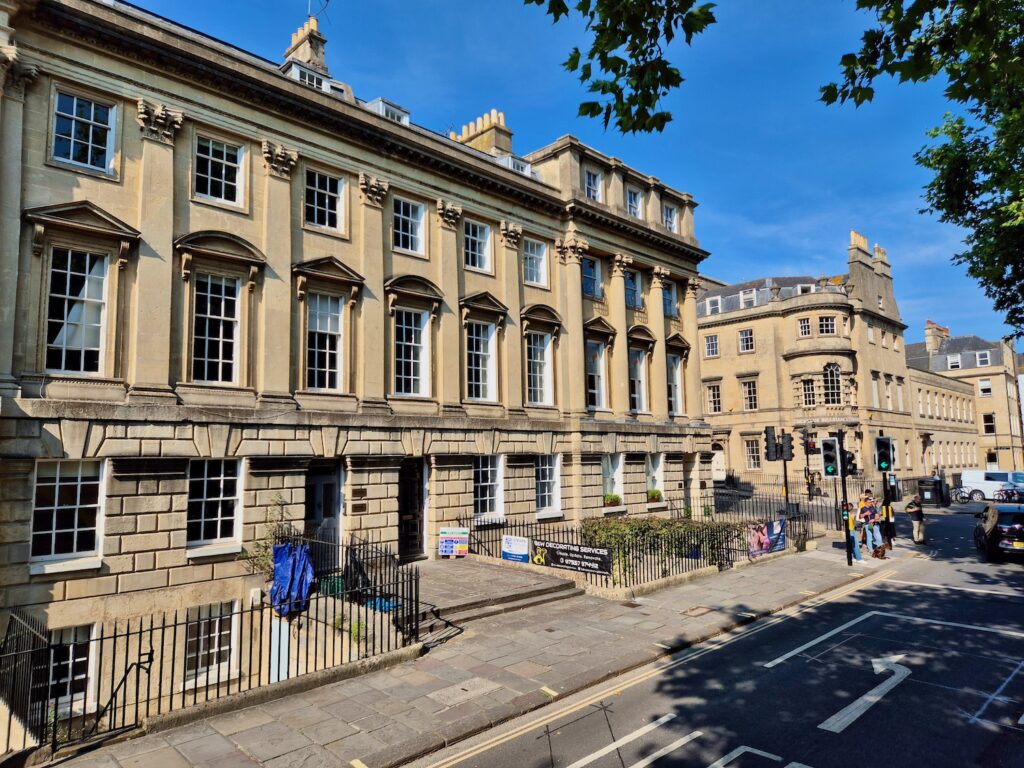 A street scene of a row of classic, light-coloured Georgian townhouses in Bath.