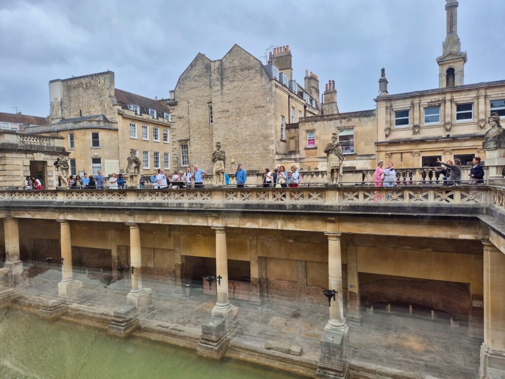 A wide view of the Roman Baths showing the main pool, columns, and a walkway with visitors.