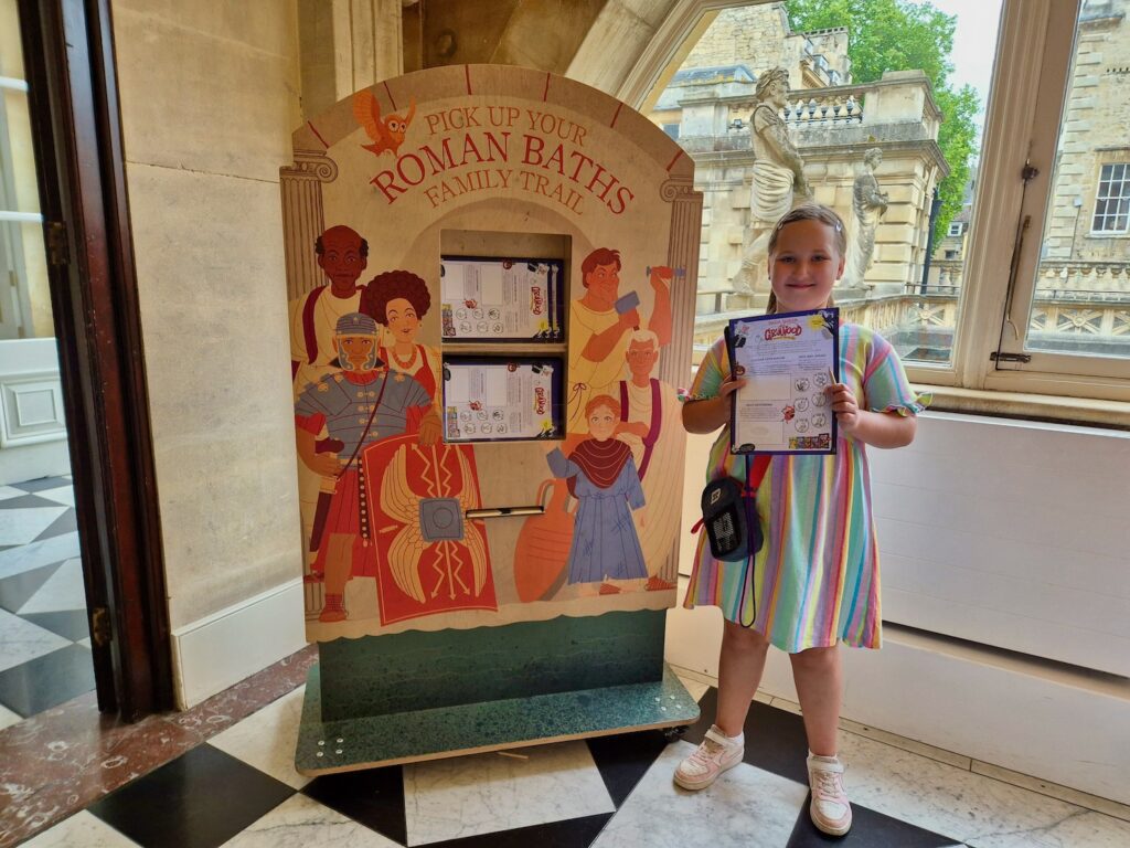 A young girl in a brightly coloured striped dress stands smiling in front of a Roman Baths "family trail" sign.