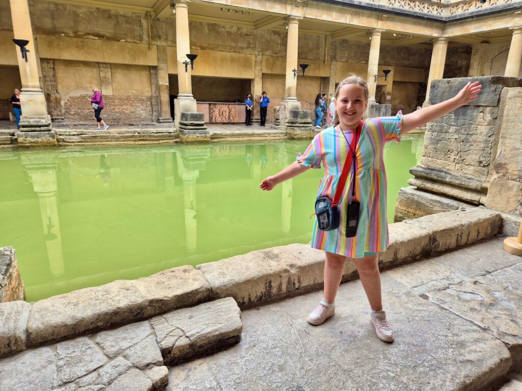 A young girl smiling with arms outspread in front of the Great Bath at the Roman Baths.