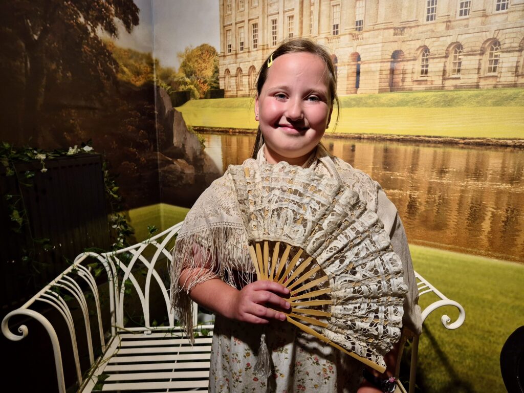 A child in a period-style dress and holding a large lace fan, posing in front of a historical backdrop at the Jane Austen Centre.