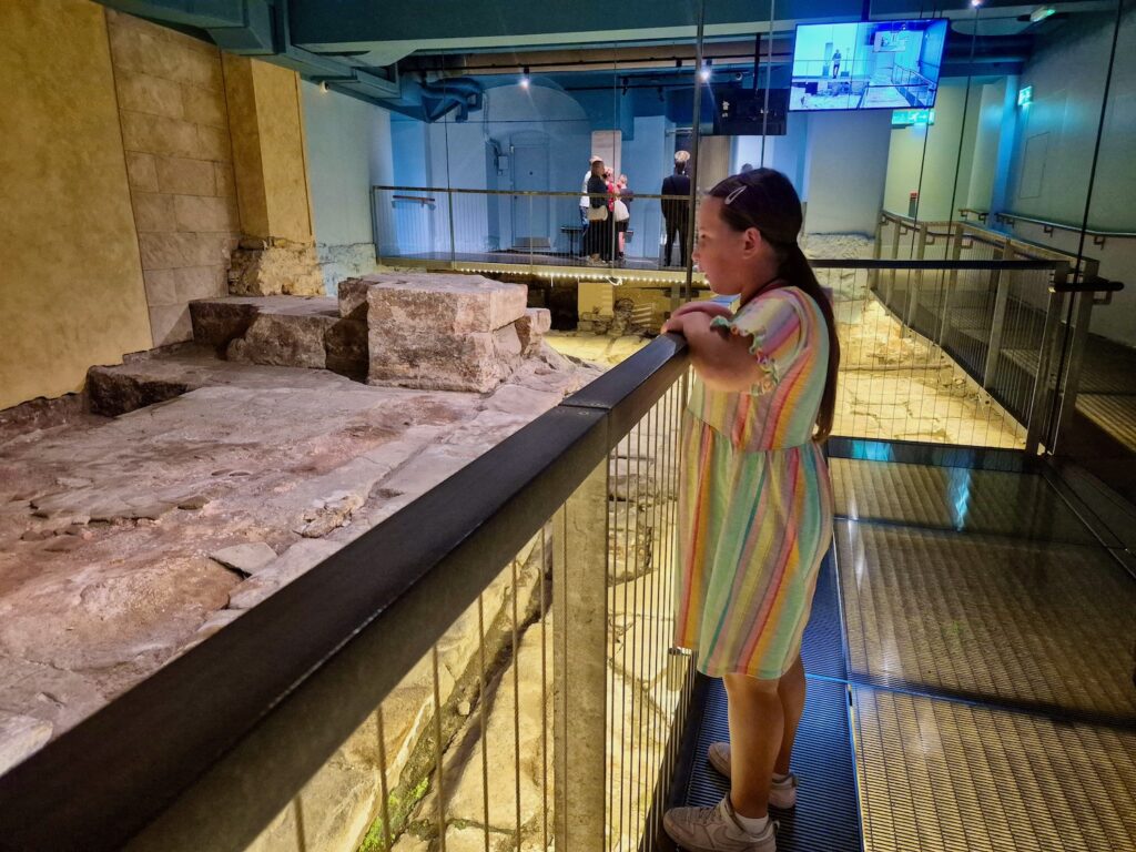 A young girl looking down at the ancient stone foundations of the Roman Baths from a raised walkway.