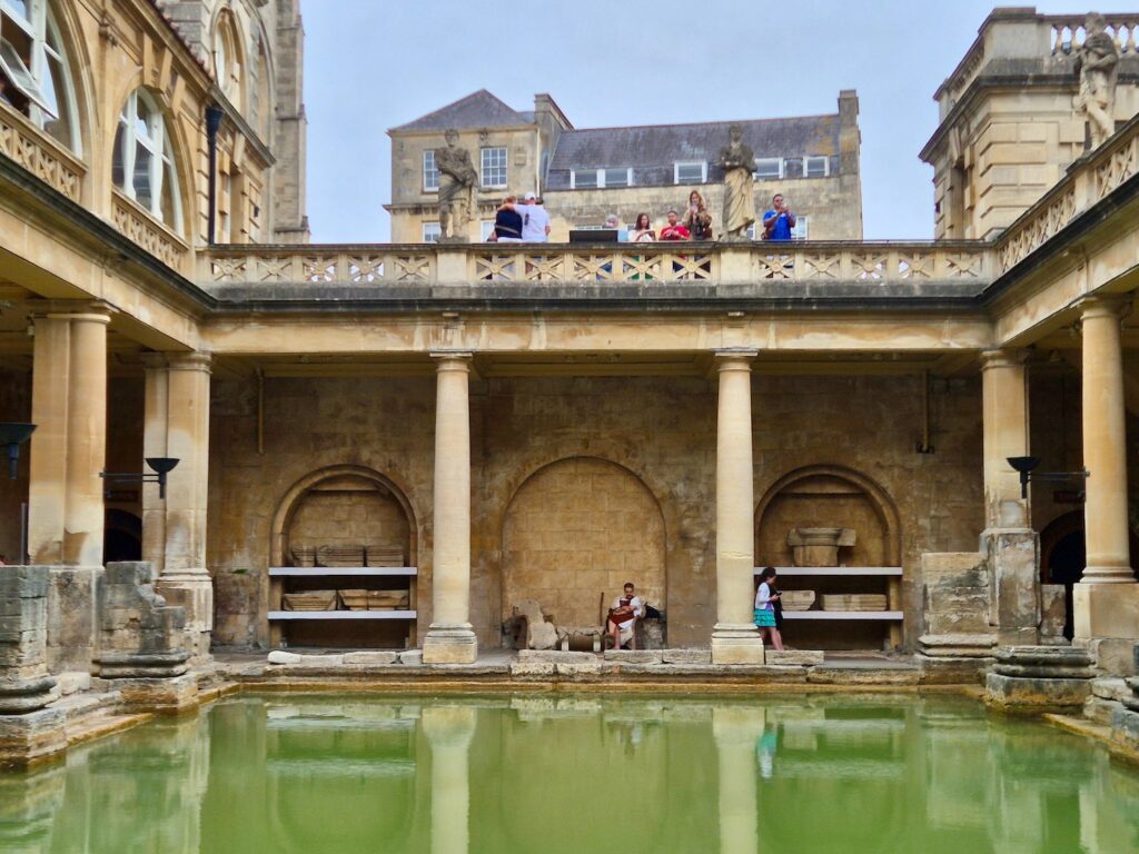 A view of the Great Bath at the Roman Baths with people on an upper balcony and a person in a tunic by the water.