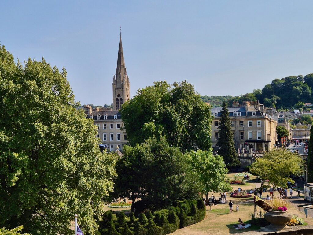 An elevated view of a landscaped park in Bath with trees, walking paths, people relaxing on the grass, and a church spire in the background.
