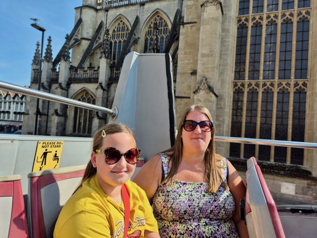 Two people seated on the top deck of a red tour bus with Bath Abbey visible behind them.